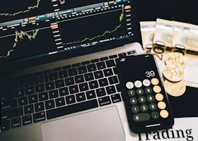 Laptop screen with trading charts, calculator app, Euros and Bitcoin coins on desk.